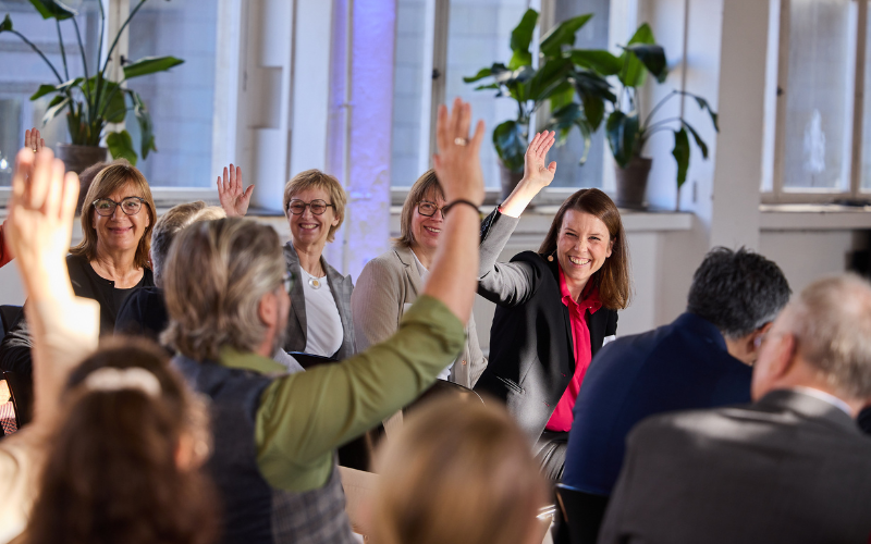 Mehrere Personen sitzen in einem Veranstaltungssaal. Einige drehen sich um und lächeln, einige heben die Hand.