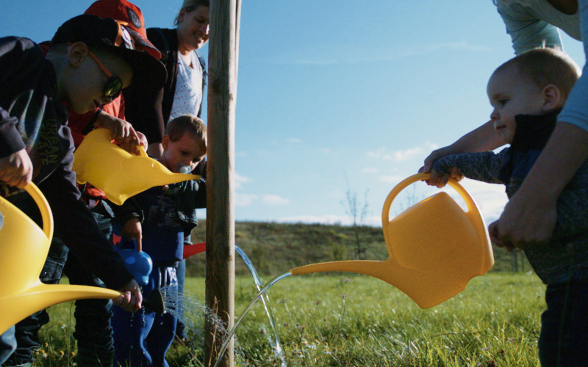 Mehrere kleine Kinder und erwachsene gießen gemeinsam einen frisch gepflanzten Baum.